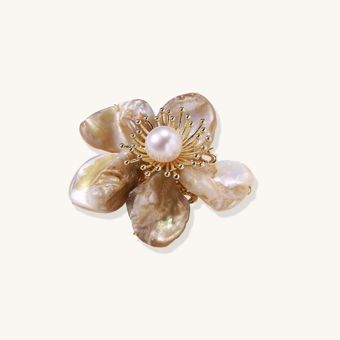 Elegant floral pin with peachy shell petals, gold radiating spokes, and white freshwater pearl center on cream background.