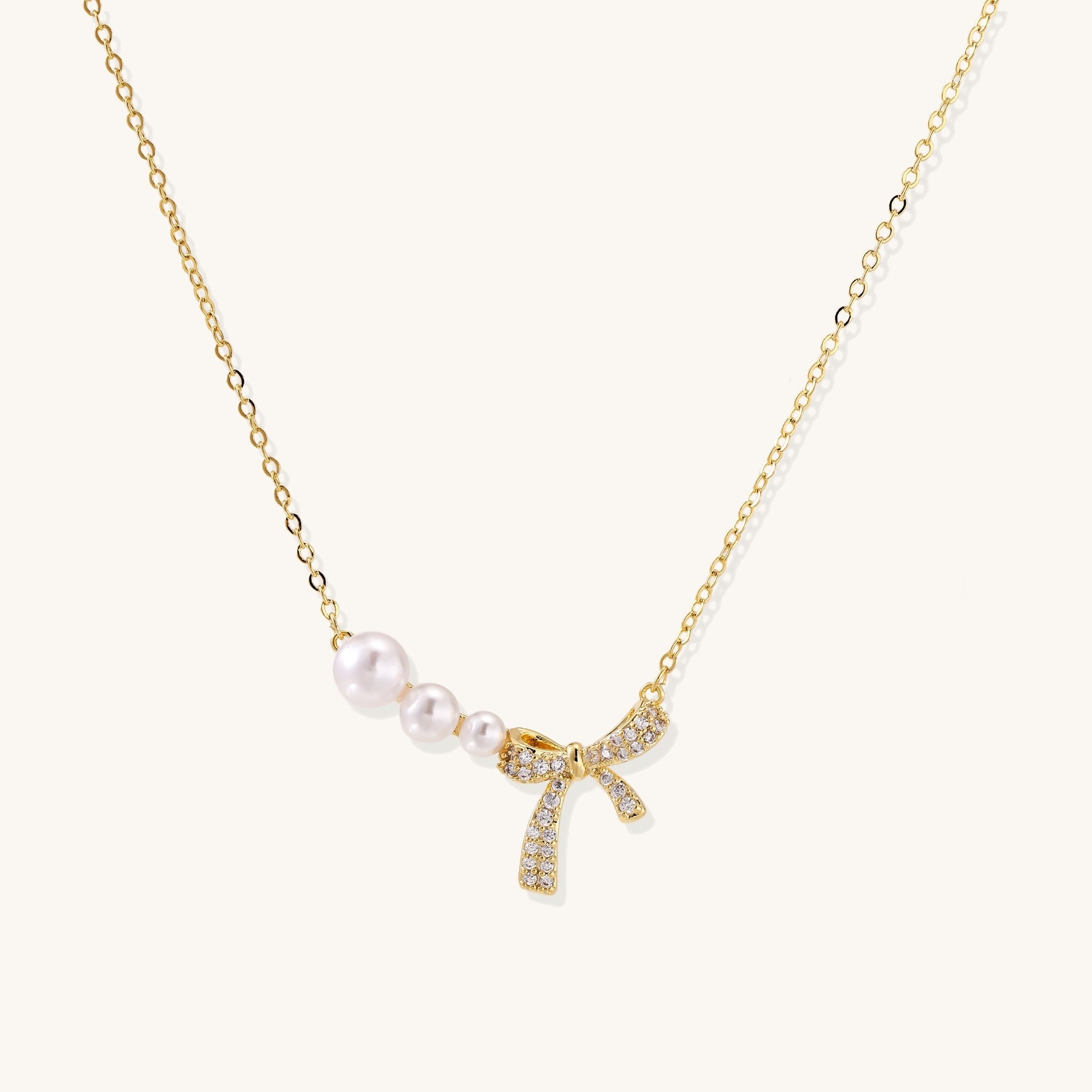 Delicate gold-plated necklace with three white pearls and sparkling bow pendant on cream background.