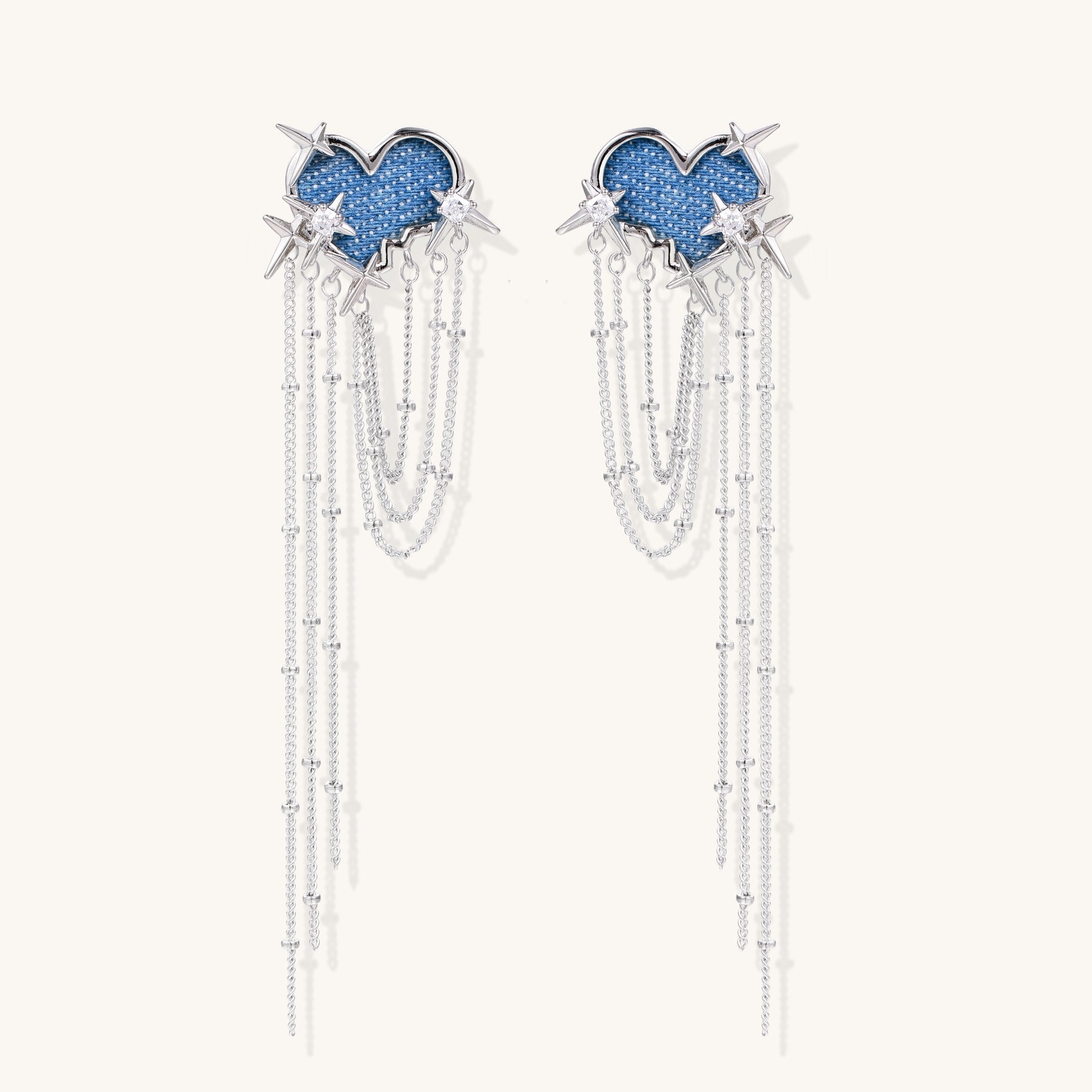 Long dangle earrings with blue denim heart shapes, silver star spikes, and multiple cascading chains on beige background.