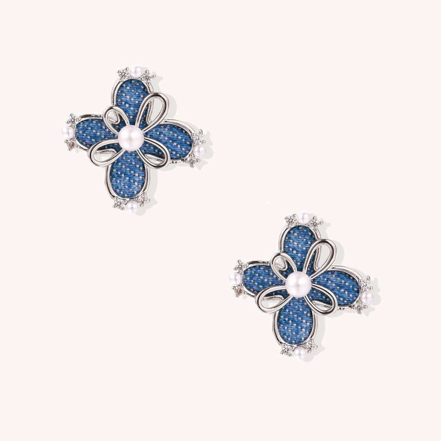 Blue denim flower stud earrings with silver framework, white pearl centers, and crystal accents on cream background.