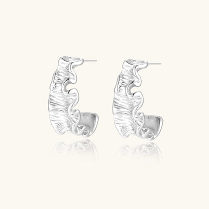 White gold plated sculptural hoop earrings with organic wavy edges and reflective metallic finish on cream background