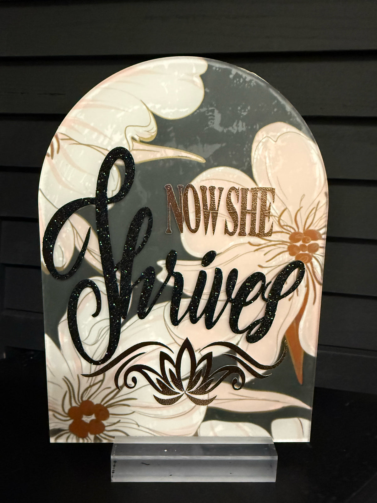 Now She Thrives arched acrylic plaque: blush-cream magnolias on charcoal, black glitter script and copper foil lotus, on a clear acrylic stand